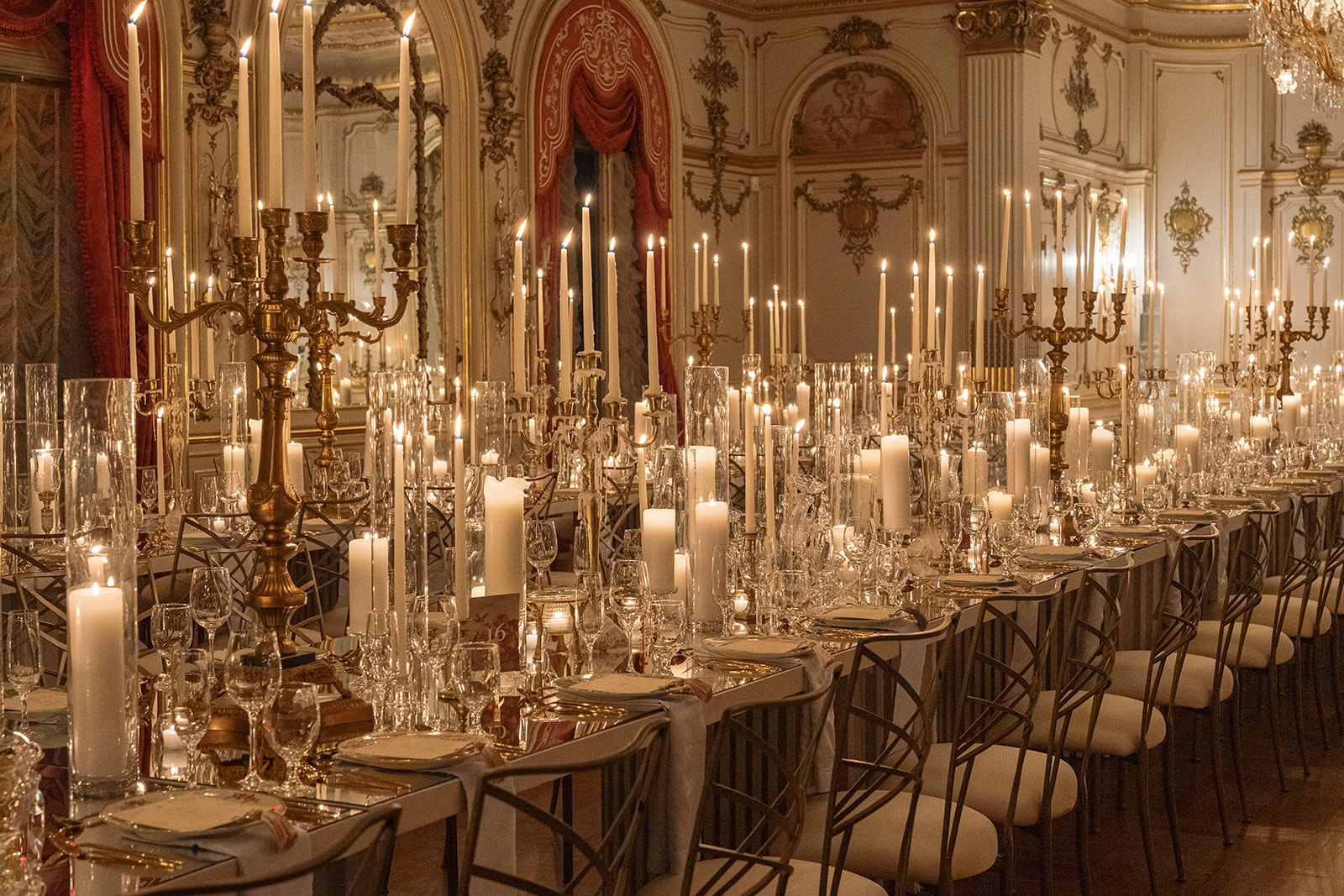 Long mirrored tablescapes and gold Fanfare chairs inside a historic candlelit ballroom wedding reception.